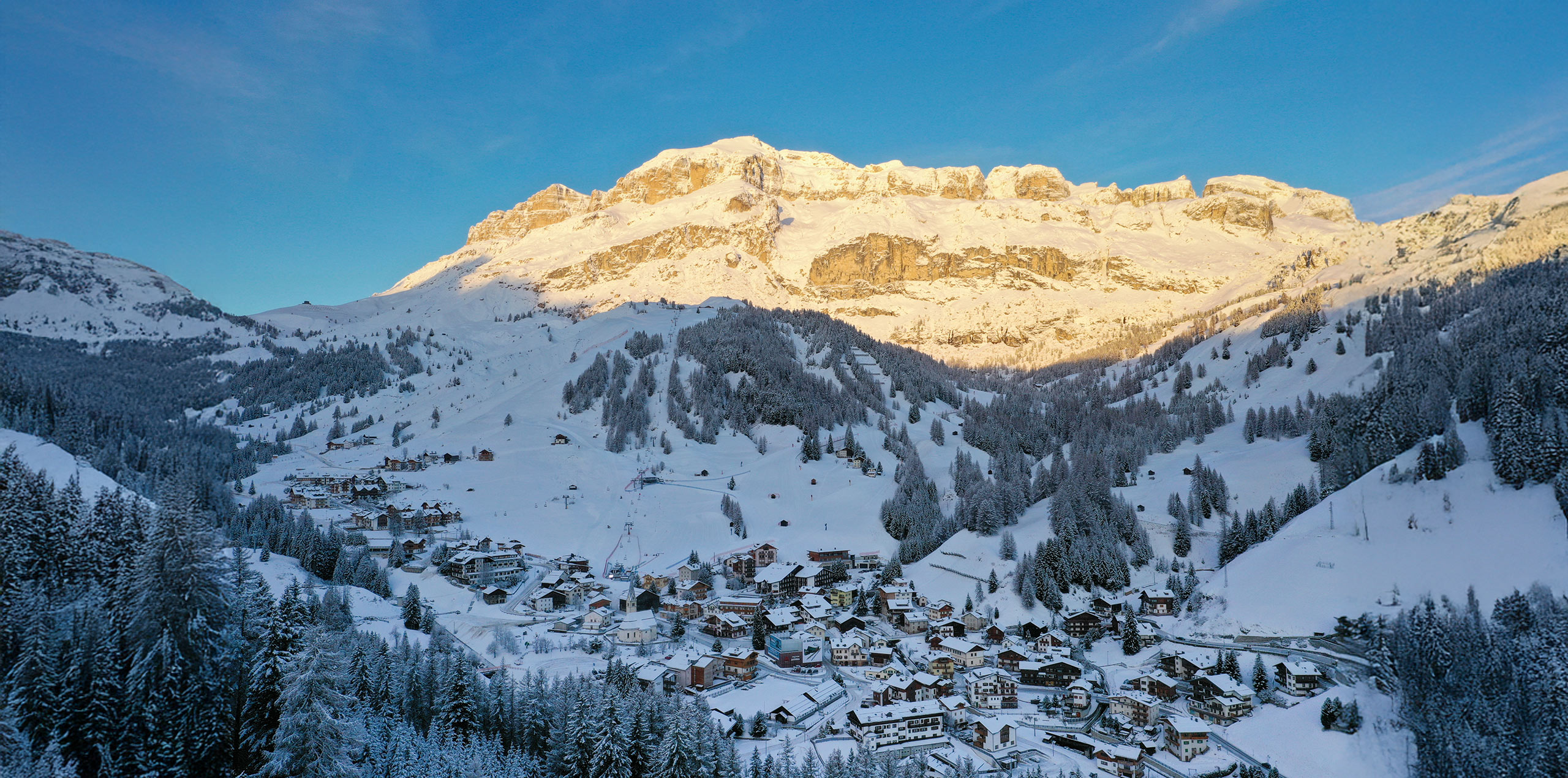 Arabba, cuore delle Dolomiti, in un mattino d'inverno. Il paese innevato è circondato da abeti e montagne illuminate dal sole.