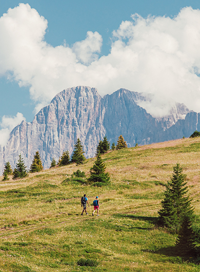 Due escursionisti camminano su un sentiero erboso in montagna, con le Dolomiti e nuvole bianche sullo sfondo.