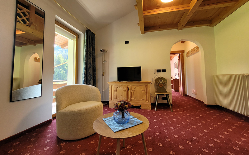 Garni Royal suite in Arabba with an armchair, small table, TV on a wooden cabinet, and a view of the Dolomites.