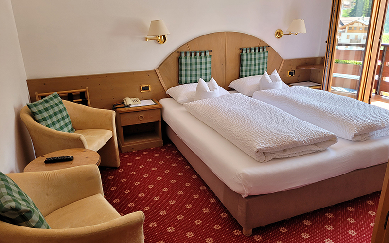 Cozy double room at Garni Royal in Arabba with single beds, wooden furnishings, armchairs, and a balcony with a view of the Dolomites.