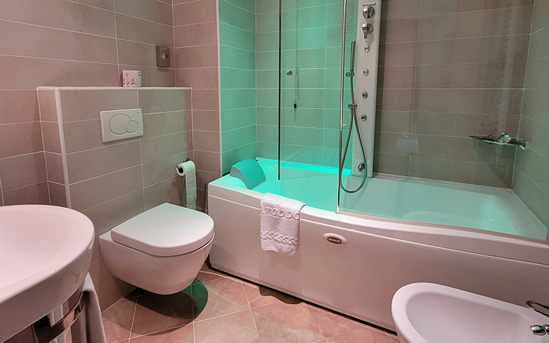 Modern bathroom with light tiles, green-lit whirlpool tub, wall-mounted shower, and wall-hung sanitary ware.