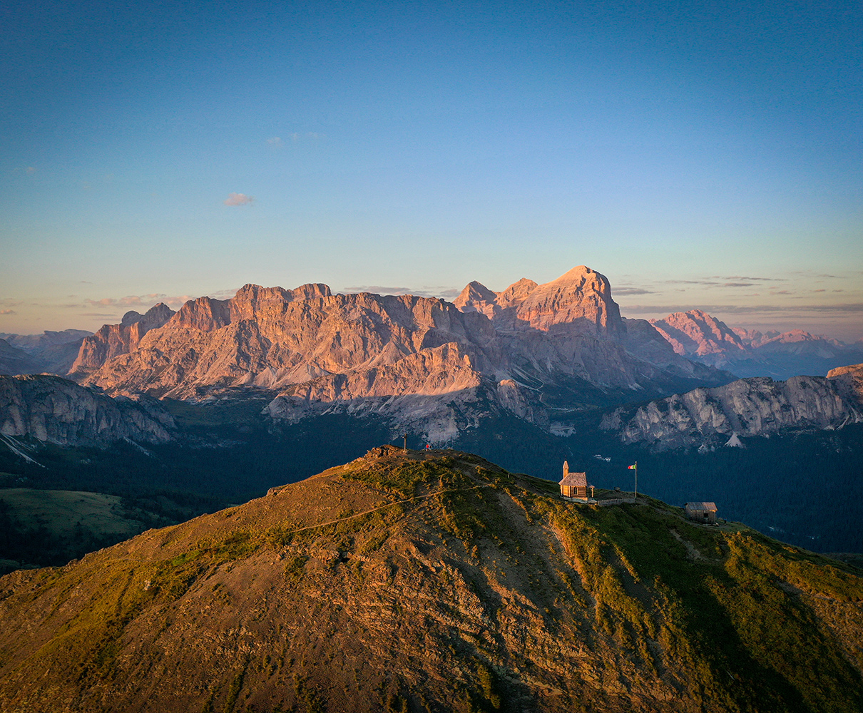 Vista panoramica delle Dolomiti al tramonto con una chiesetta e un rifugio su un crinale, ideale per le vacanze a Arabba.