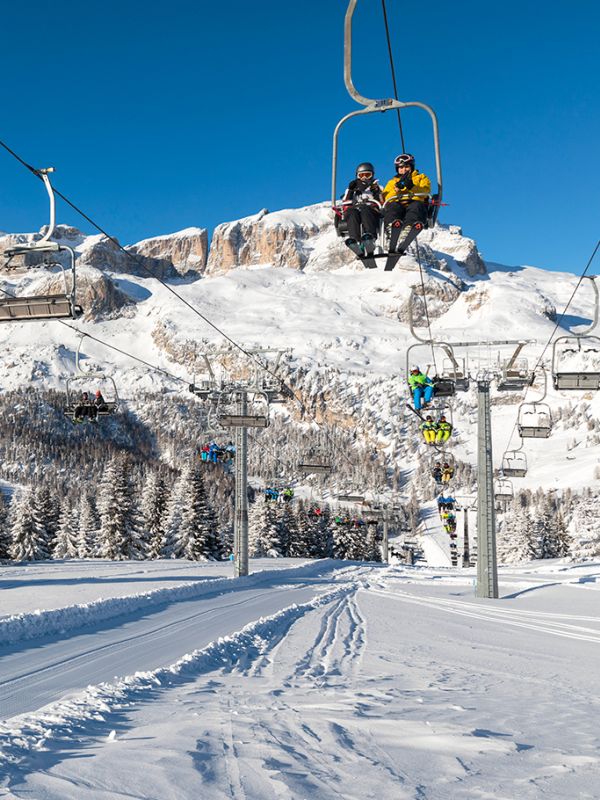 Sciatori salgono con la seggiovia sulle Dolomiti innevate di Arabba, con piste e boschi sotto un cielo azzurro.